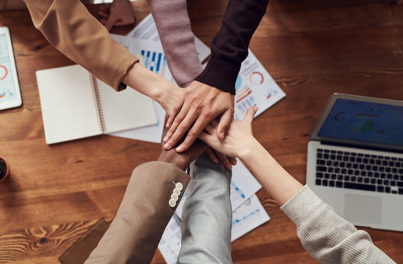 6 people overlapping their hands in a "let's do this!" type of style. There is a laptop, a few notebooks, some colorful charts and graphs on a wooden table visible.
